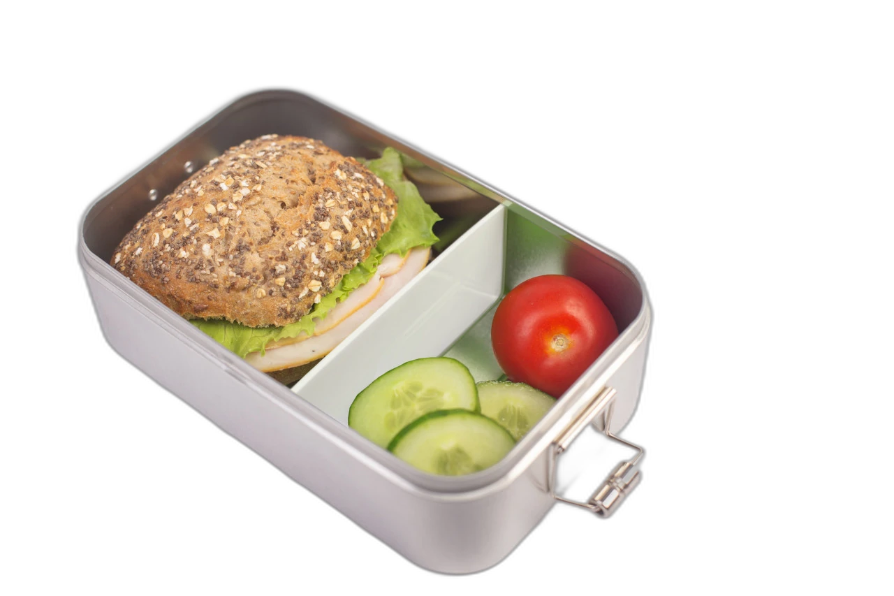 Boîte à pain rectangulaire couleur argent avec cloison blanche insérée, remplie d'un petit pain aux graines avec salade ainsi que des tranches de tomate et concombre, photographié sur fond blanc.