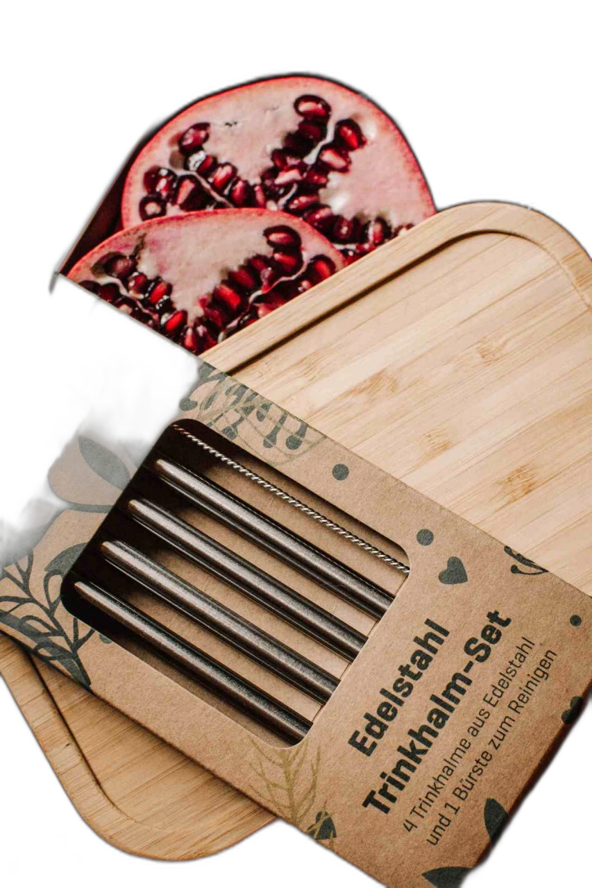 Ensemble de pailles à boire durables en acier inoxydable dans un emballage en carton sur un couvercle en bambou, à l'arrière-plan une boîte métallique ouverte contenant des moitiés de grenade frais et des myrtilles sur une table en bois.