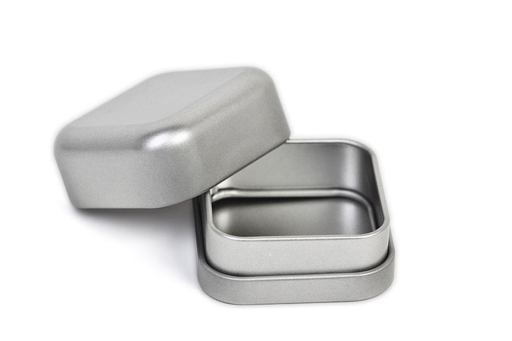 Petite boîte métallique carrée ouverte en métal couleur argent, couvercle légèrement retiré à côté du corps, photographiée sur fond blanc.