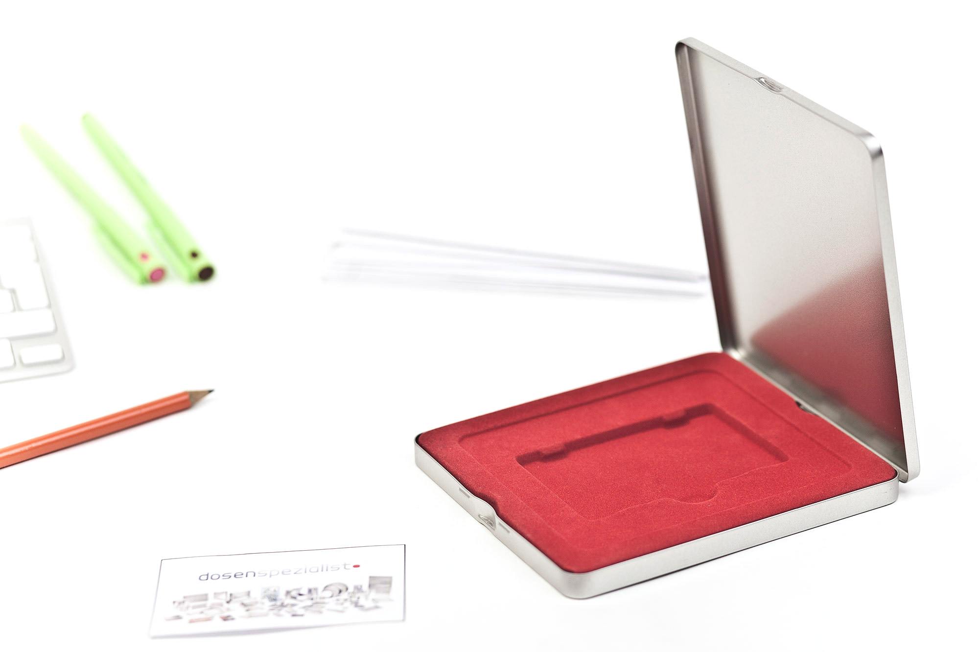 Boîte métallique angulaire ouverte en métal couleur argent avec couvercle à charnière, intérieur avec insert en mousse rouge pour accueillir une carte plastique. Photographiée sur fond blanc avec articles de bureau en arrière-plan.