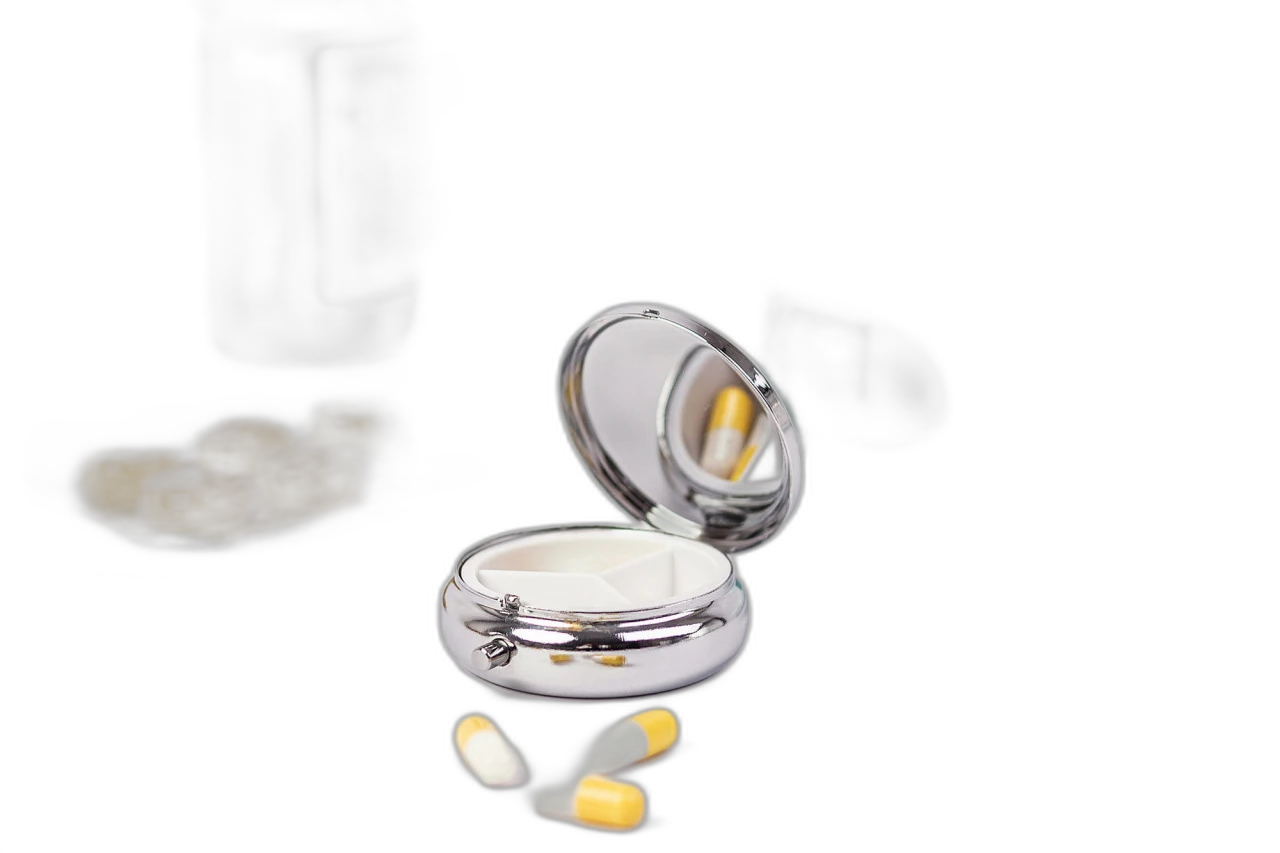 Boîte à pilules ronde ouverte en métal couleur argent avec trois compartiments intérieurs et miroir intégré, photographiée avec comprimés et médicaments en arrière-plan.