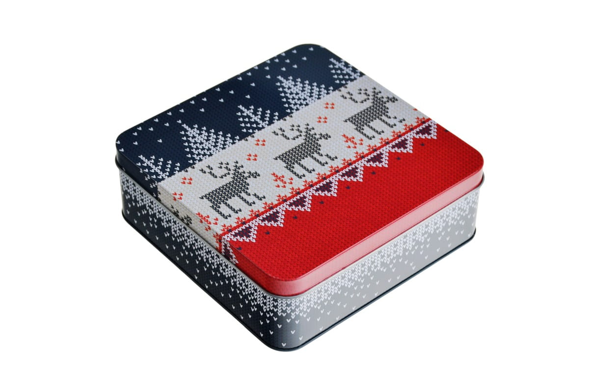 Boîte en fer blanc rectangulaire fermée avec motif tricoté de Noël en bleu, rouge et blanc, avec sapins stylisés et rennes représentés. Le couvercle a une bordure rouge et est photographié sur fond blanc.