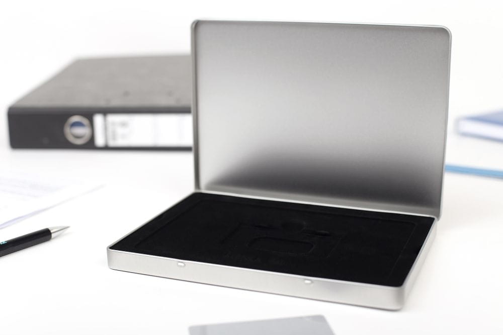 Boîte à bon d'achat rectangulaire ouverte en métal couleur argent au format A5, avec insert en mousse noire pour un positionnement sécurisé, photographiée sur fond clair avec articles de bureau en arrière-plan.