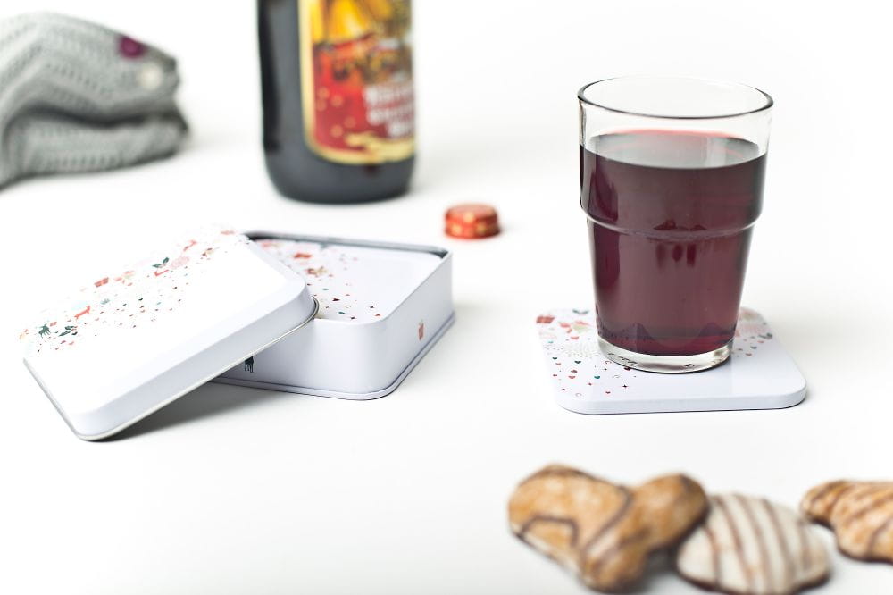 Un verre contenant une boisson rouge posé sur un dessous de verre blanc métallique, à côté d'une boîte ouverte au design festif avec quelques biscuits. Le fond est neutre.