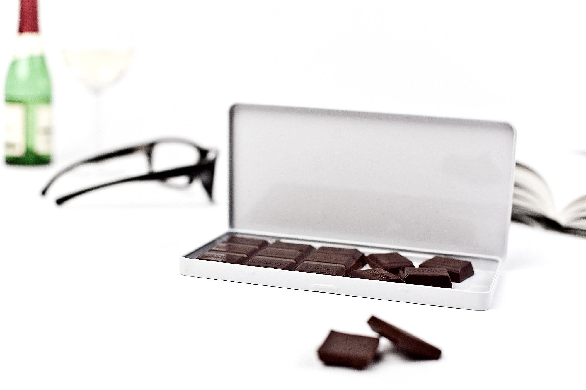 Boîte métallique blanche pour chocolat ouverte avec morceaux de chocolat, à côté d'un verre de vin, d'une bouteille et de lunettes, sur fond blanc.