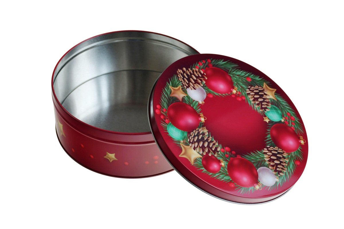 Boîte métallique ronde ouverte avec motif de Noël, couvercle avec boules rouges, étoiles, branches de sapin et pommes de pin posé à côté, photographiée sur fond blanc.