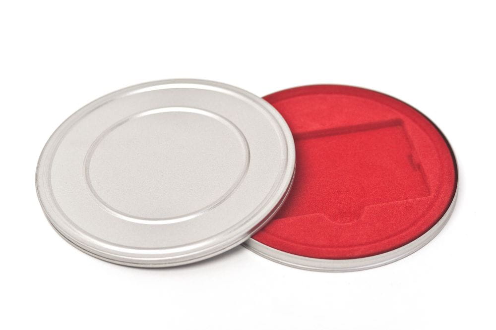 Boîte métallique ronde ouverte en métal couleur argent avec couvercle retiré, intérieur avec doublure en velours rouge et compartiment pour cartes, photographiée sur fond blanc.