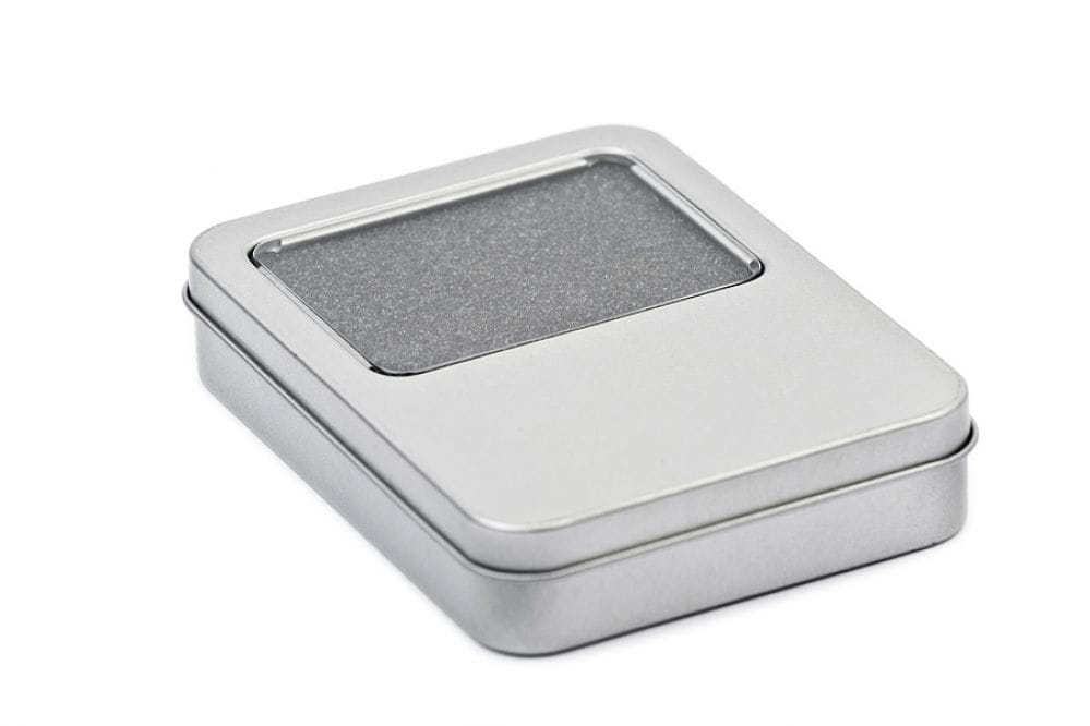 Boîte métallique rectangulaire fermée en argent avec fenêtre transparente dans le couvercle, photographiée sur fond blanc neutre.