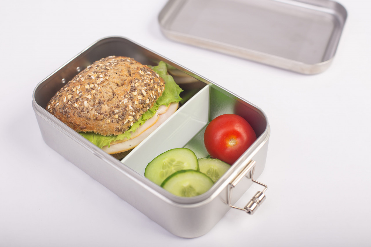 Boîte à lunch en acier inoxydable Classic Maxi avec cloison blanche insérée, remplie de petit pain garni, tomate et tranches de concombre ; couvercle en arrière-plan, fond neutre.